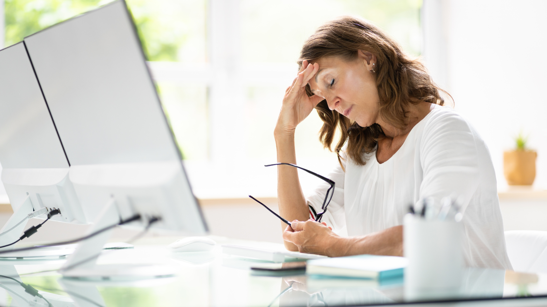 Fatigued woman at a glass desk with monitors, holding her glasses with a hand to her forehead