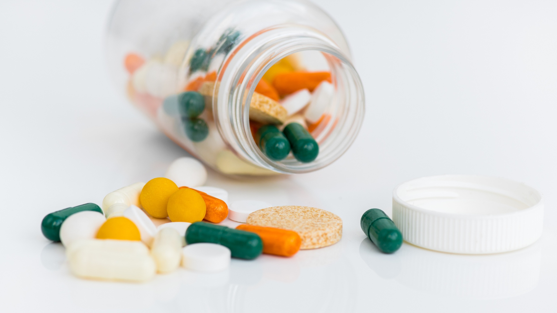 Colorful vitamin pills and capsules spilling from a clear bottle on a white surface